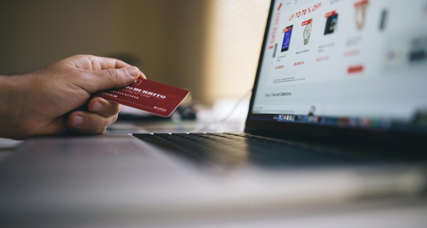 Hand holding a red credit card in front of a laptop with an online shopping website displayed.